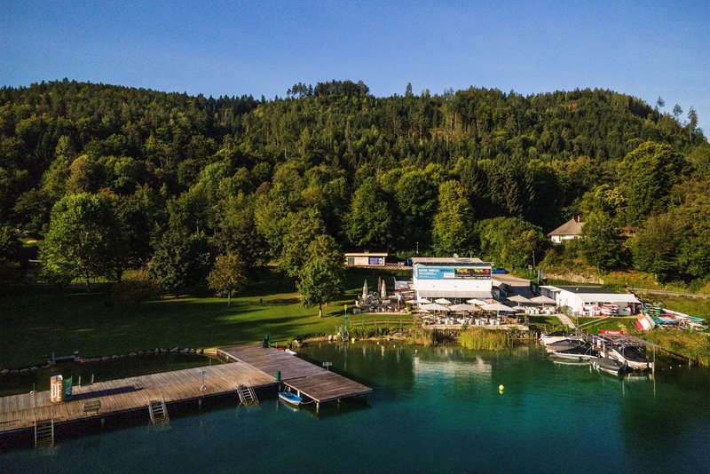 Luftaufnahme des Strandbads Maiernigg: Badesteg und Bootsanleger im türkisen Wörthersee, Restaurant-Terrasse und Liegewiese am bewaldeten Ufer