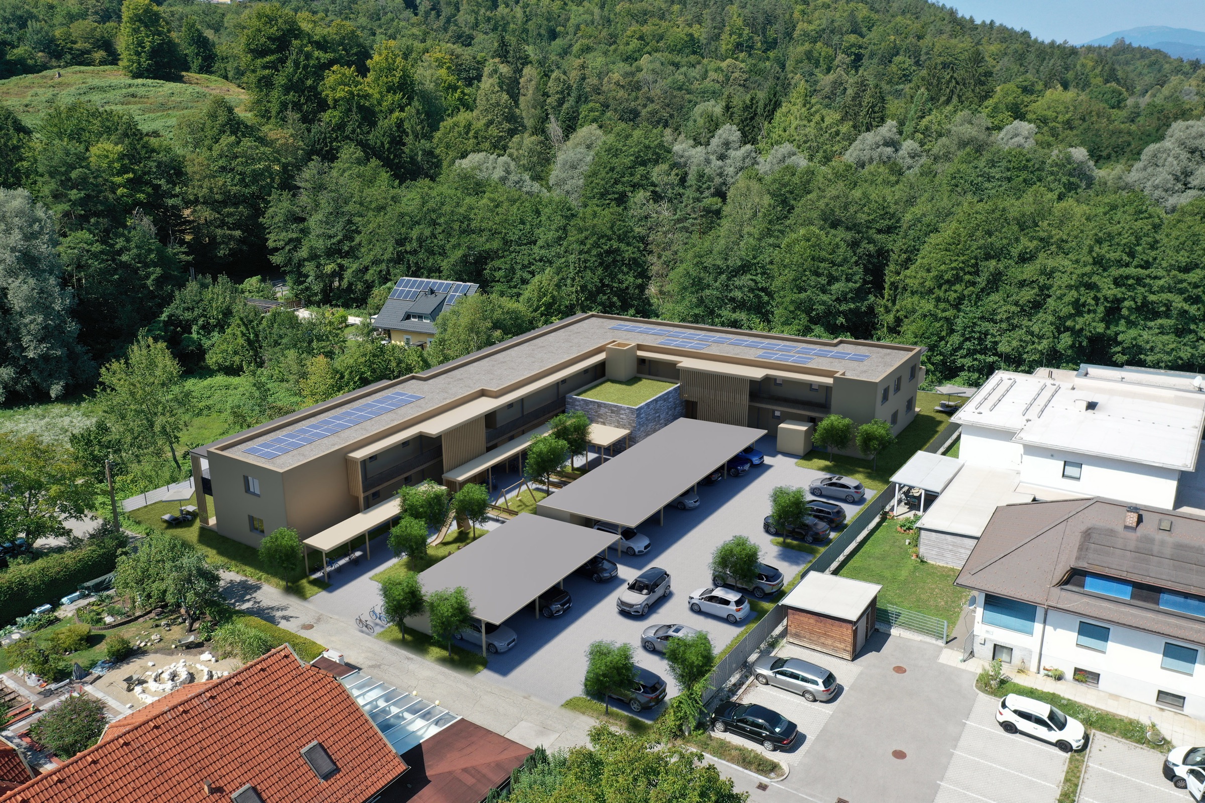 Luftaufnahme des L-förmigen Neubaus mit Solarpaneelen auf dem Flachdach, überdachten Stellplätzen, Spielplatz und angrenzenden Einfamilienhäusern am bewaldeten Hang