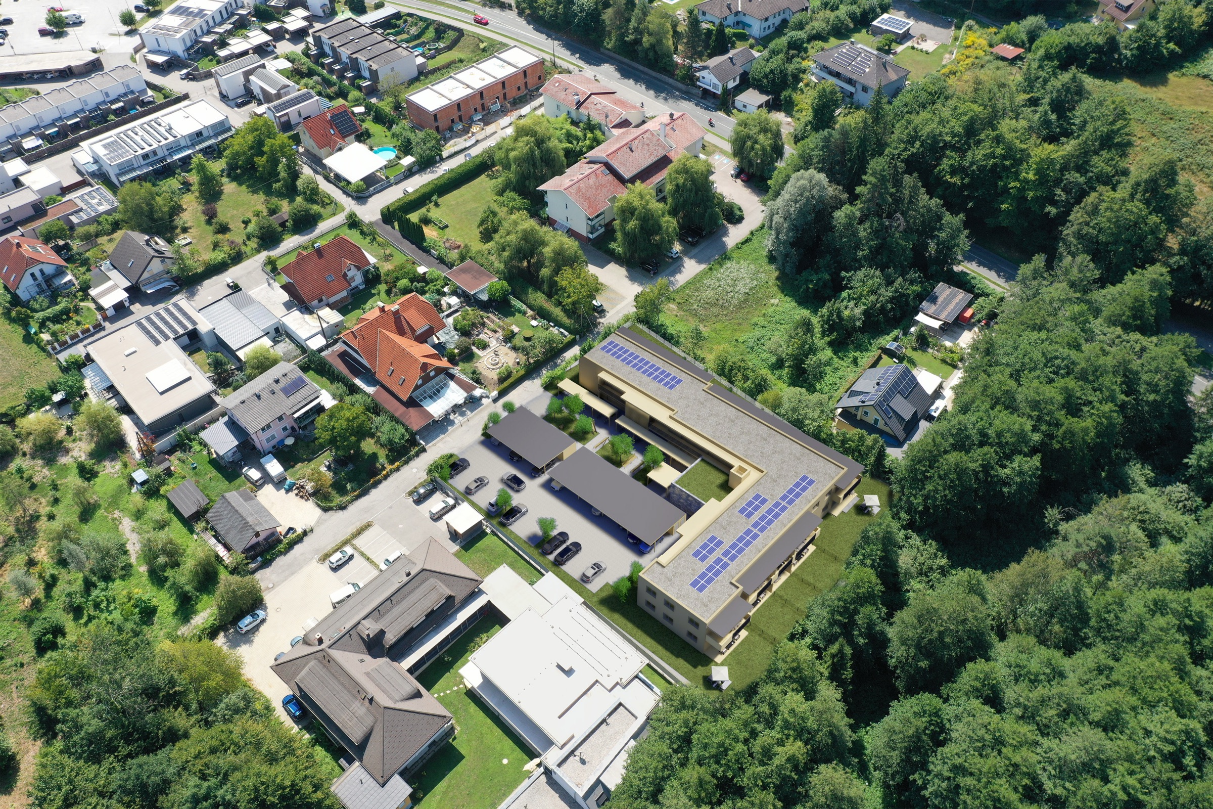 Luftaufnahme aus größerer Distanz: der Neubau eingebettet in ein bestehendes Wohngebiet mit Einfamilienhäusern, Straße und angrenzender Waldfläche
