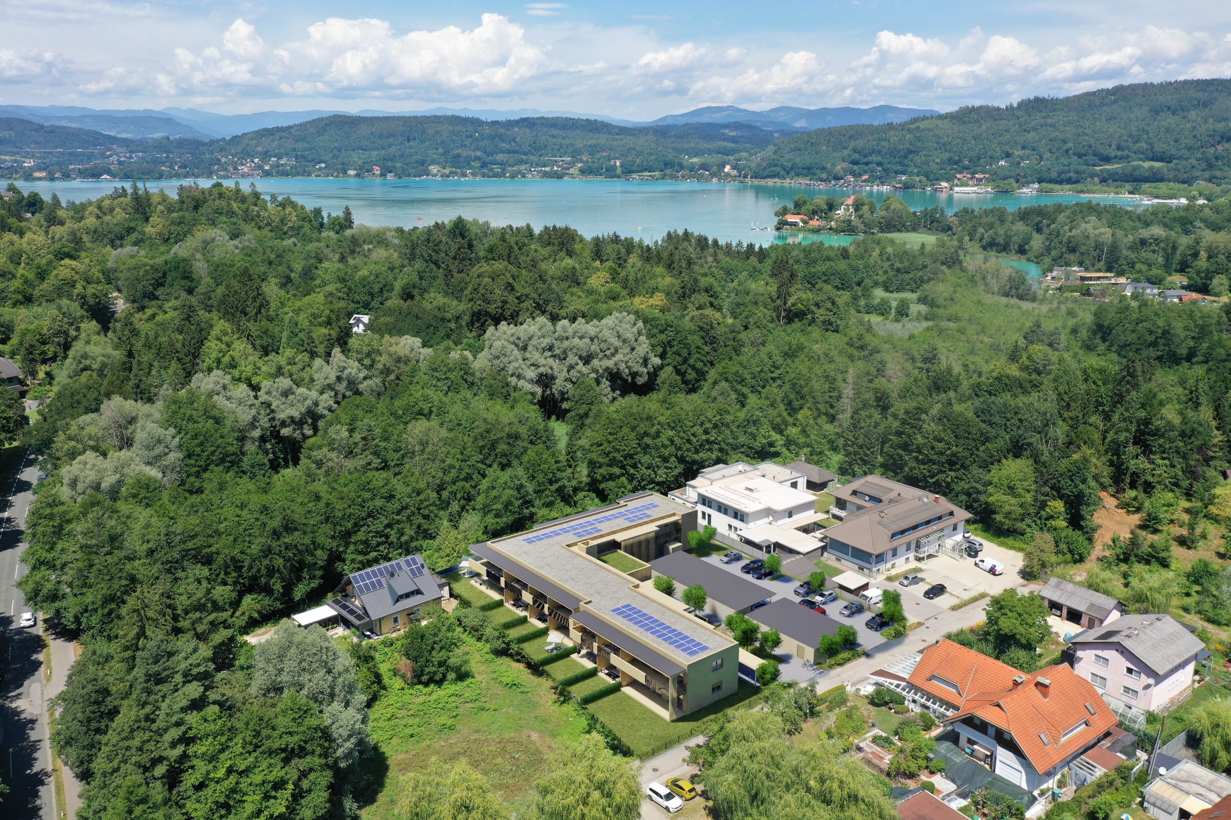 Luftaufnahme der Projektumgebung: der Neubau von SÜD:See und ein angrenzendes Bestandsgebäude vor Waldflächen, dahinter der türkisfarbene See und die Bergkette