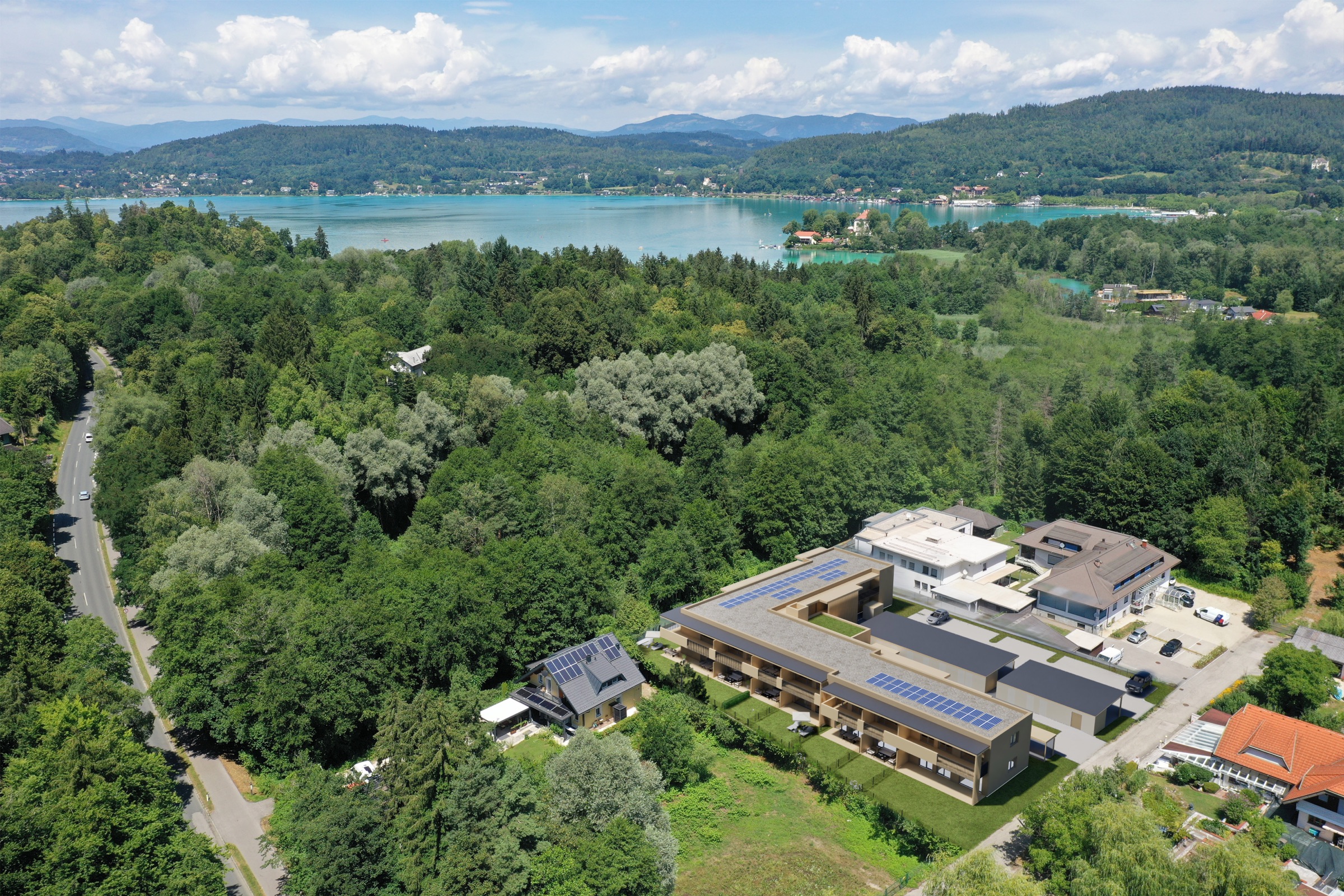 Luftaufnahme der Umgebung: der Neubau von SÜD:See mit angrenzendem Bestandsgebäude, davor ein Einfamilienhaus mit Photovoltaikanlage, dahinter der See und die Bergkette
